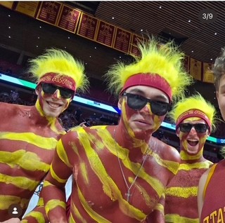 Adam and two friends in cardinal and gold body paint and wigs, cheering in the student section at a game.